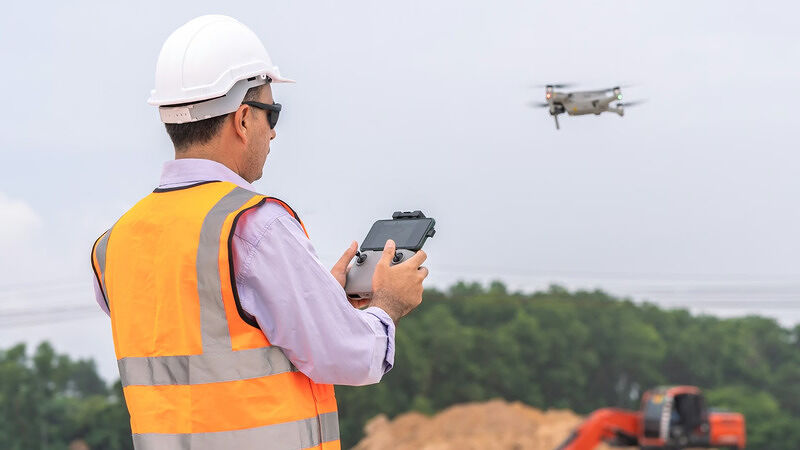 Person wearing a hard hat and high-visibility vest operating a drone remote outdoors over a field
