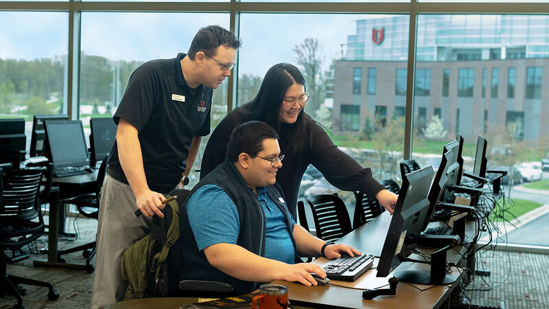 Instructor leaning over to guide a student working at a computer station in a classroom