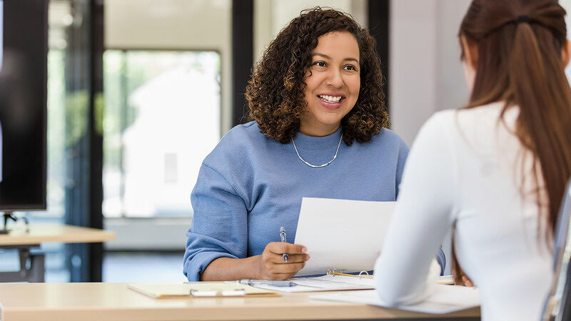 Professional woman in a blue top sitting across a table, smiling while reviewing paper documents during a meeting