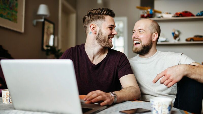 Two men sitting together at a desk, smiling while looking at a laptop computer screen