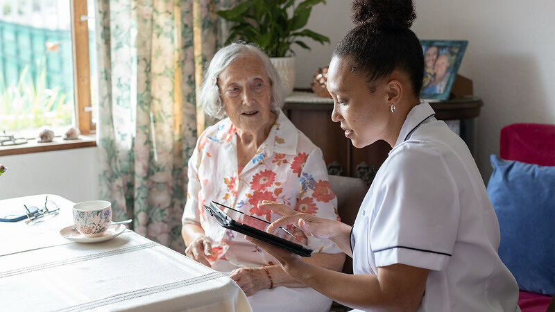 Healthcare worker sitting at a table outdoors assisting an elderly woman with a digital tablet