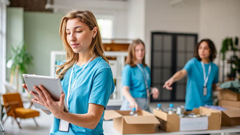 Three volunteers in light blue shirts working together indoors one using a tablet while two others sort items into cardboard boxes on a table in a bright room with shelves plants and large windows