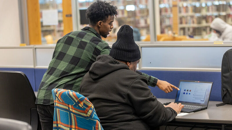 Two people at a desk in a library one sitting and using a laptop while another stands beside them pointing at the screen with bookshelves and another person in the background