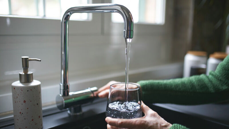 A person in a green sweater filling a clear glass with water from a chrome faucet in a kitchen with a soap dispenser and containers near a window in the background
