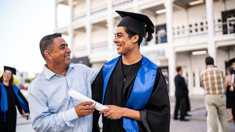 Graduate in a navy blue cap and gown smiling and holding a diploma outdoors while chatting with an older man