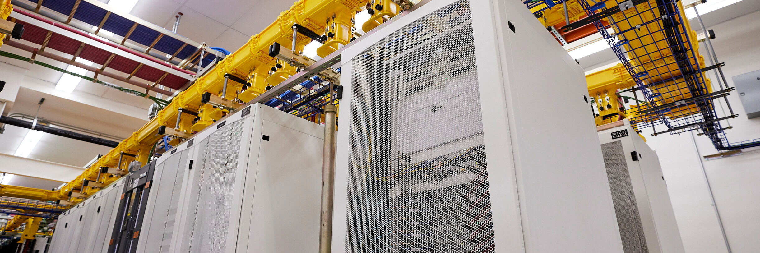 A brightly lit server room with tall white racks filled with hardware and overhead trays carrying yellow and blue cables